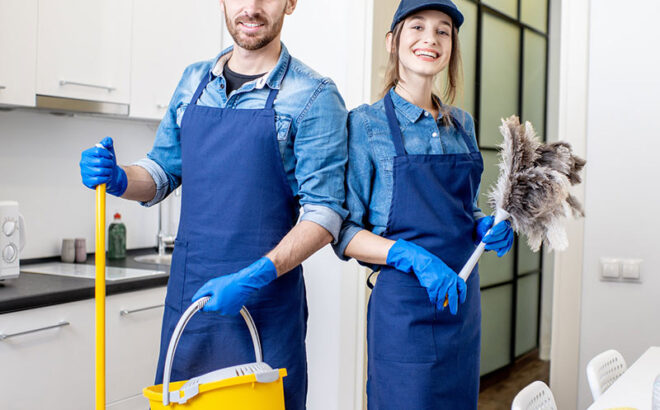 Portrait of a couple as a professional cleaners in uniform standing together with cleaning tools indoors